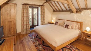 A bedroom with a wooden ceiling, a bed with pillows, a nightstand with a lamp, a wooden wardrobe, and a window with curtains. - The Old Stables - UKC4806 (Bishops Nympton)