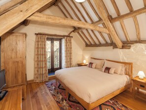 A bedroom with a wooden ceiling, a bed with pillows, a nightstand with a lamp, a wooden wardrobe, and a window with curtains. - The Old Stables - UKC4806 (Bishops Nympton)