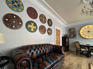 Una sala de estar con un sofá de cuero, arte decorativo en la pared, un reloj de pared grande y un comedor con mesa y sillas.