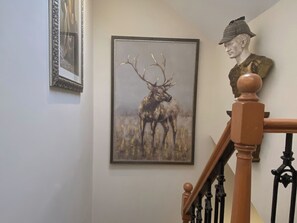 A staircase with a wooden handrail and black balusters, a framed painting of a deer, and a bust of a man wearing a hat. - There’s a copper bath in the bedroom - Enough said. (Berwick-on-Tweed)