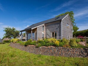 A two-story house with a stone retaining wall, a sloped roof, and a garden with various plants. - Orchard Cottage - UKC4805 (Bishops Nympton)