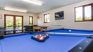 Taula de bitlles amb pilotes, taula de futbolí i taula de billar en una habitació amb finestres i un radiador.