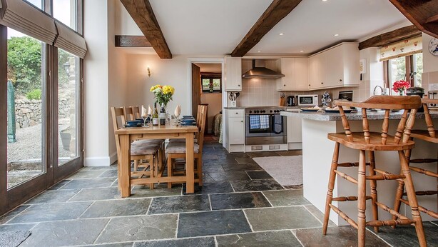 A modern kitchen with stone flooring, wooden beams, and a dining area with a table set for four. - Barn Cottage - UKC4804 (Bishops Nympton)