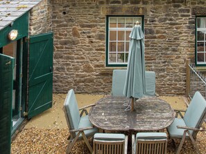 A rustic stone building with a round wooden table and four chairs, an umbrella, and a green door. - The Old Workshop - UKC4838 (New Mills)