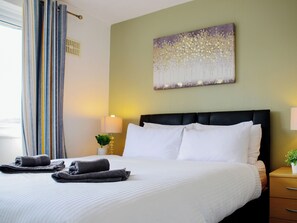 A hotel room with a bed, a nightstand, a lamp, a painting on the wall, and a window with curtains.