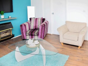 A living room with a glass coffee table, striped armchair, beige sofa, and a fireplace with logs. - St Mary's Avenue House (Stanwell)