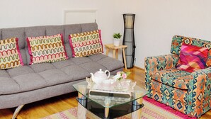 Un salon avec un canapé gris, des coussins aux motifs colorés, une table basse en verre, un fauteuil à motifs, une petite table d’appoint en bois et un grand lampadaire.
