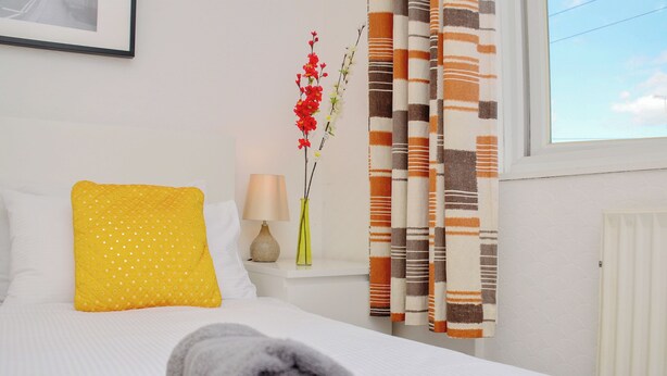 Un lit bien fait, avec une serviette enroulée, un coussin jaune, un vase de fleurs et des rideaux à rayures.