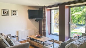 Een moderne woonkamer met een bank, een salontafel, een aan de muur bevestigde televisie en een raam met uitzicht op de omgeving.