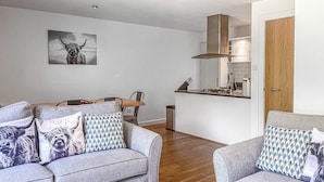 Een moderne woonkamer met grijze banken en gekleurde kussens, een houten eettafel en een keuken met een fornuis en afzuigkap.