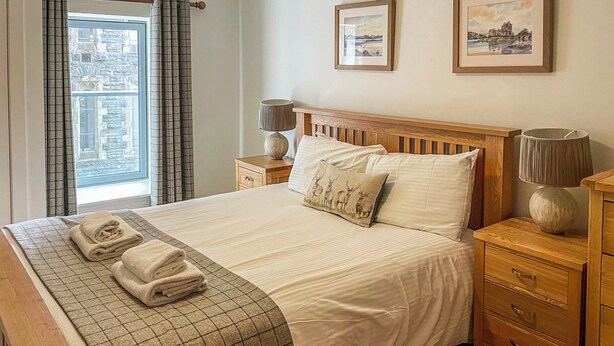 A neatly made bed with a checkered bedspread, two pillows, and folded towels. There are two bedside tables with lamps and a wooden nightstand. - Raven Wing 11 - Kintail Apartment - UKC4883 (Fort Augustus)