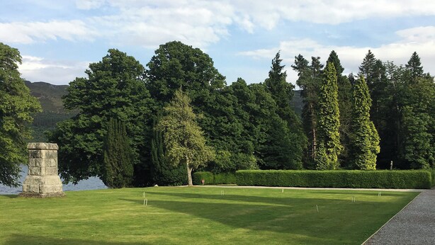 A well-maintained lawn with a stone monument, neatly trimmed hedges, and tall trees in the background. - Moat House 6 - UKC4889 (Fort Augustus)