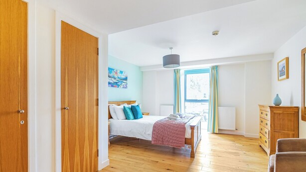 Een slaapkamer met houten vloer, een bed met wit en blauw beddengoed, een raam met gordijnen, een radiator en een houten dressoir.