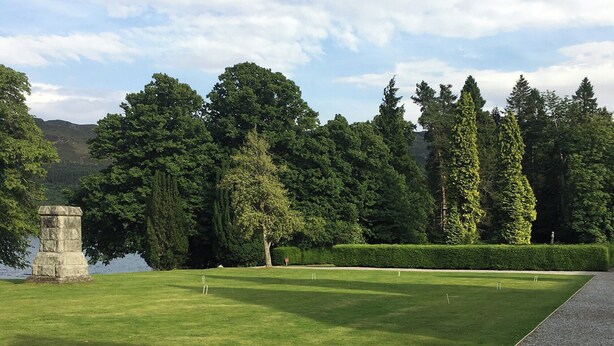 A well-maintained golf course with a stone monument, neatly trimmed hedges, and a variety of trees in the background. - Abbey Garden - UKC4894 (Fort Augustus)