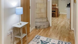 A hallway with a wooden floor, a patterned rug, a lamp on a small table, and a stone pillar. - Abbey Garden - UKC4894 (Fort Augustus)