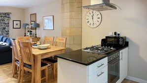 A compact kitchen with a gas stove, microwave, and oven, a dining table with chairs, and a clock on the wall. - Abbey Garden - UKC4894 (Fort Augustus)