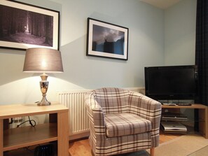 A living room with a plaid armchair, a lamp, a television, and framed pictures on the wall. - Raven Wing 14 - Glenwood - UKC4891 (Fort Augustus)