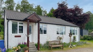 Une petite maison de plain-pied, à l’extérieur blanc et au toit foncé. On y trouve un banc en bois à l’extérieur, un petit jardin avec différentes plantes et une allée gravillonnée.