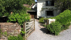 Un bâtiment traditionnel de couleur blanche, avec un chemin de gravier menant à un portail en bois, le tout entouré d’une végétation luxuriante.