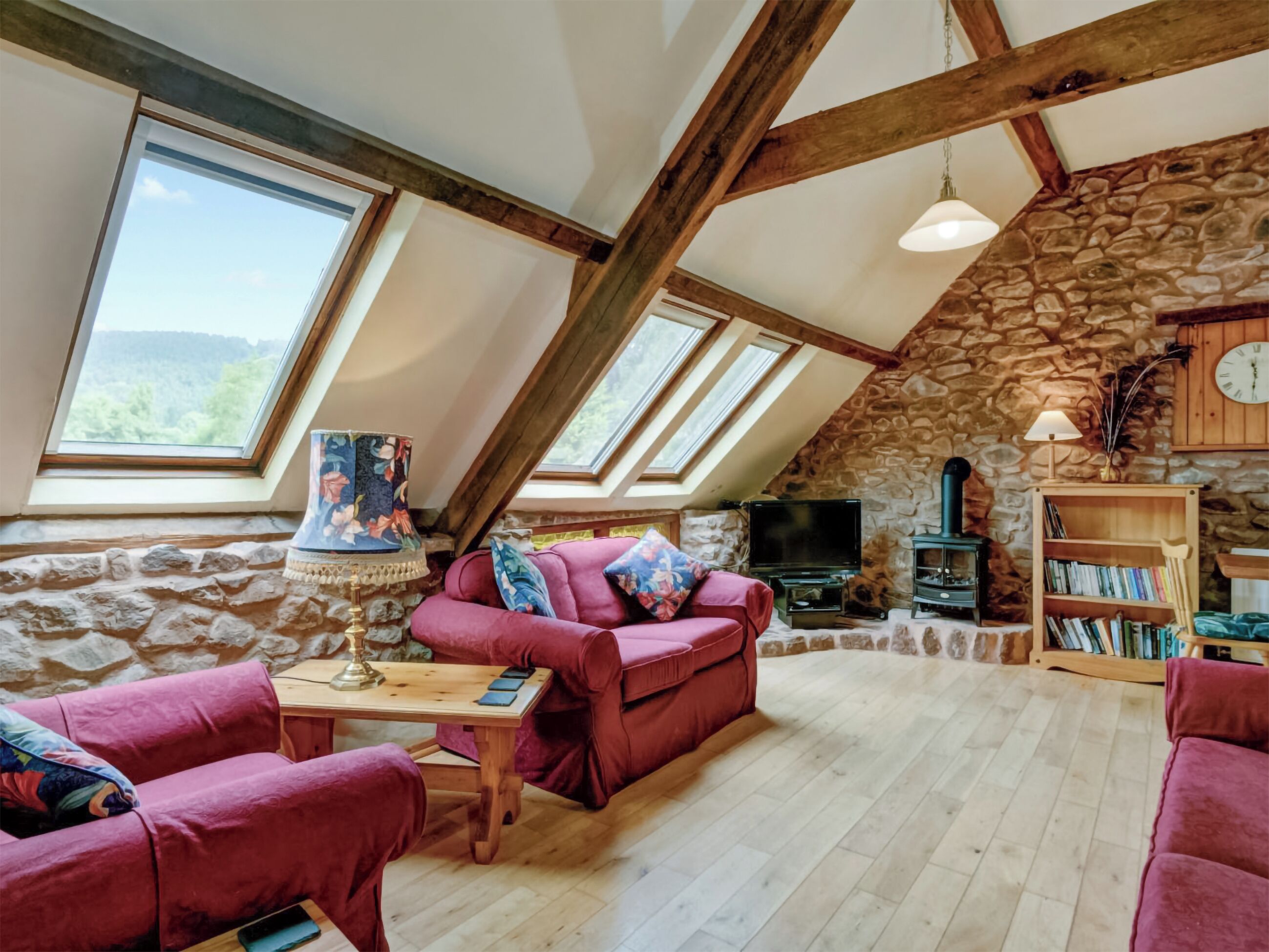 A cozy loft with a stone wall, wooden beams, a red sofa, a TV, and a bookshelf.