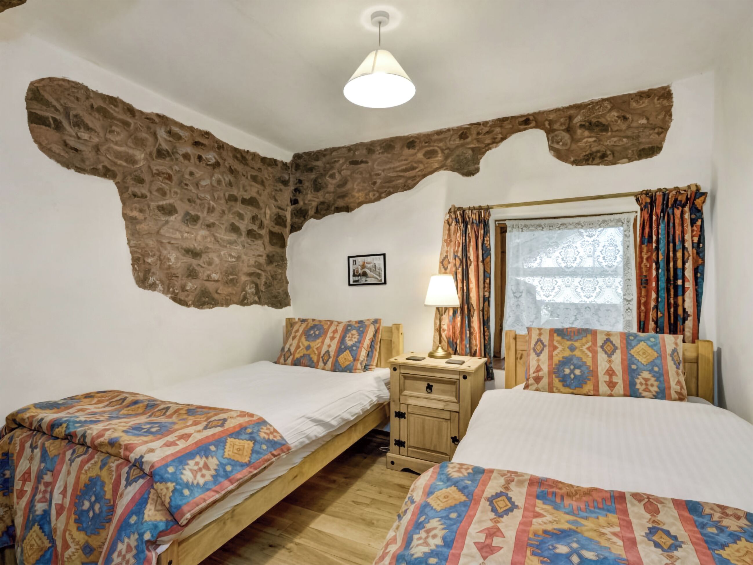 A room with two beds, a wooden nightstand, a lamp, a patterned window curtain, and a stone wall.