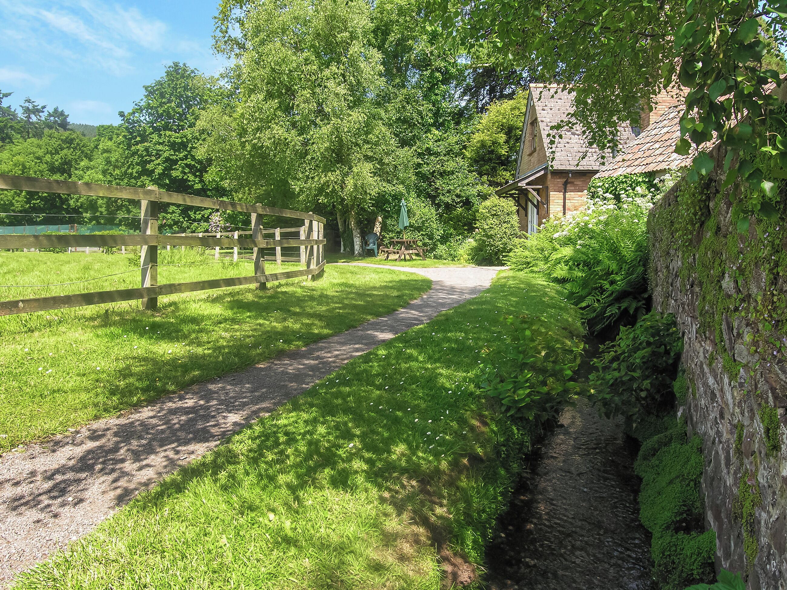 A narrow gravel path leading to a rustic building with a tiled roof, surrounded by lush greenery and a wooden fence.