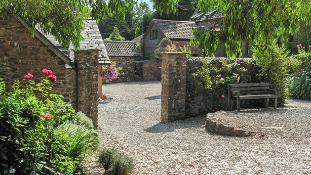 A stone building with a gravel path, a wooden bench, and lush greenery. - 2 bedroom accommodation in Timberscombe, near Minehead (Timberscombe, near Minehead)