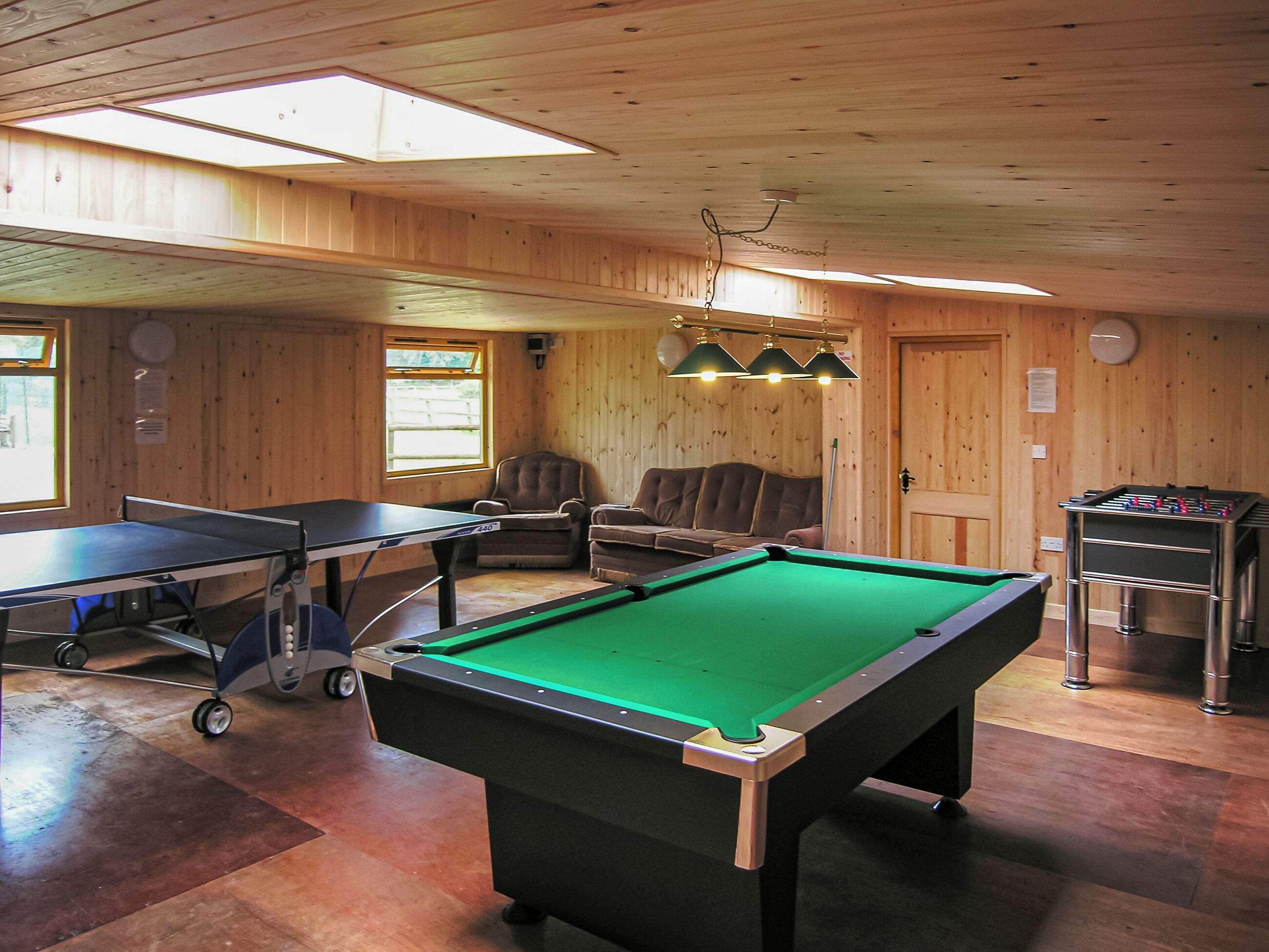 A pool table, a foosball table, and a ping pong table in a room with wooden walls and ceiling, large windows, and a ceiling fan.