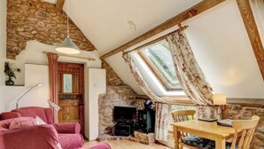 A cozy living room with a stone wall, a wooden ceiling, a TV, a fireplace, a dining table with chairs, and a pink sofa. - 1 bedroom accommodation in Timberscombe, near Minehead (Timberscombe, near Minehead)