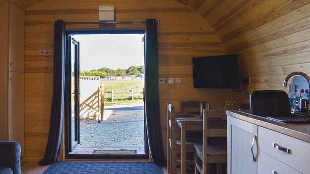 Een houten interieur met een deur naar buiten, een zithoek met stoelen en een tafel, een tv aan de muur en een kleine kitchenette met een spoelbak en aanrecht.
