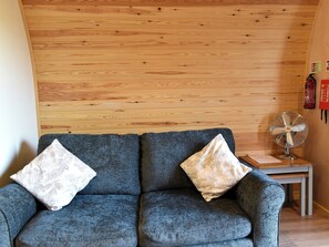 A room with a wooden ceiling, a grey sofa with floral cushions, and a standing fan on a small table. - 1 bedroom accommodation in Ferndown (Ferndown)