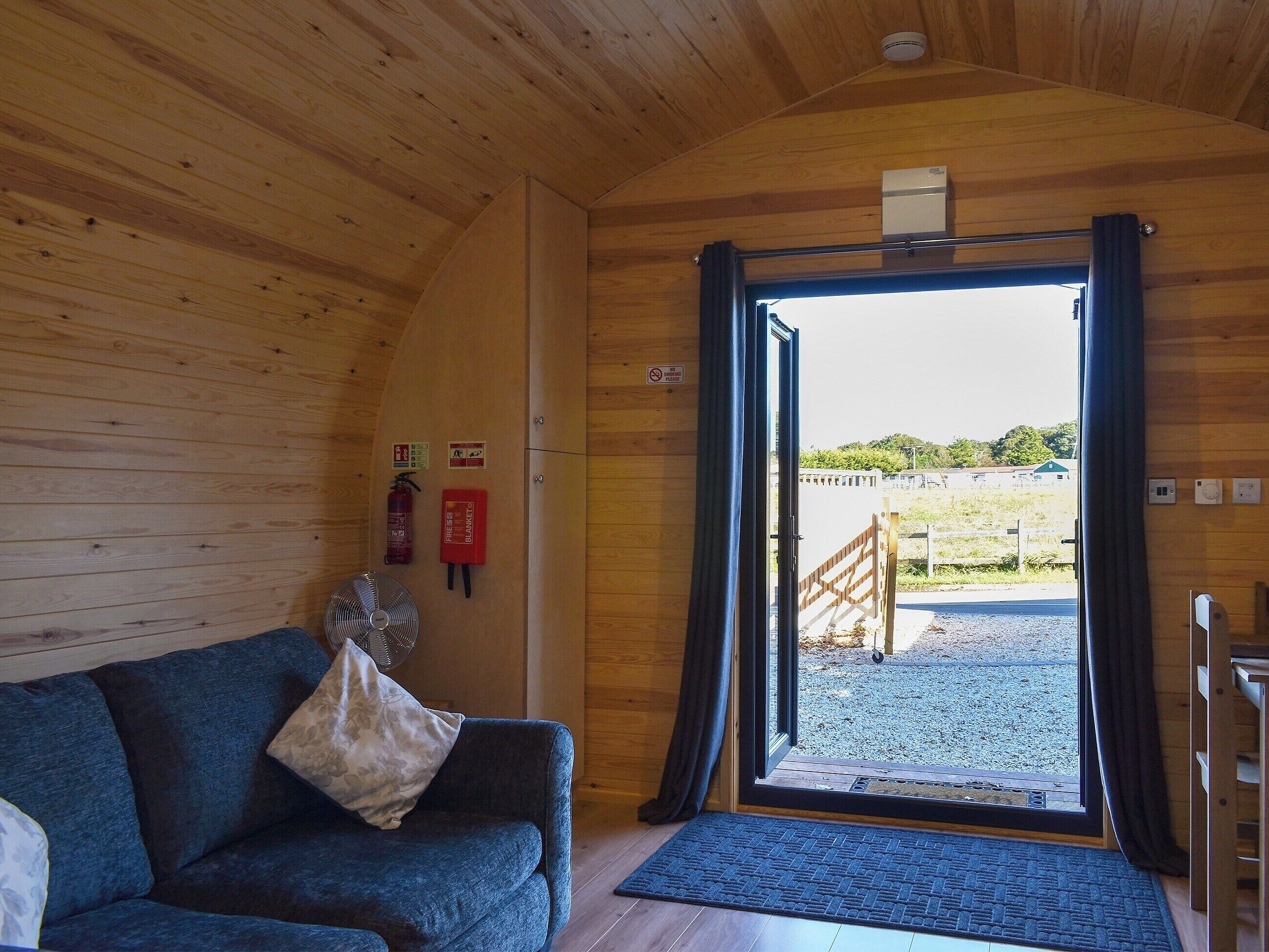 Une pièce chaleureuse avec un plafond en bois, un canapé bleu avec un coussin, un extincteur et une porte donnant sur l’extérieur.