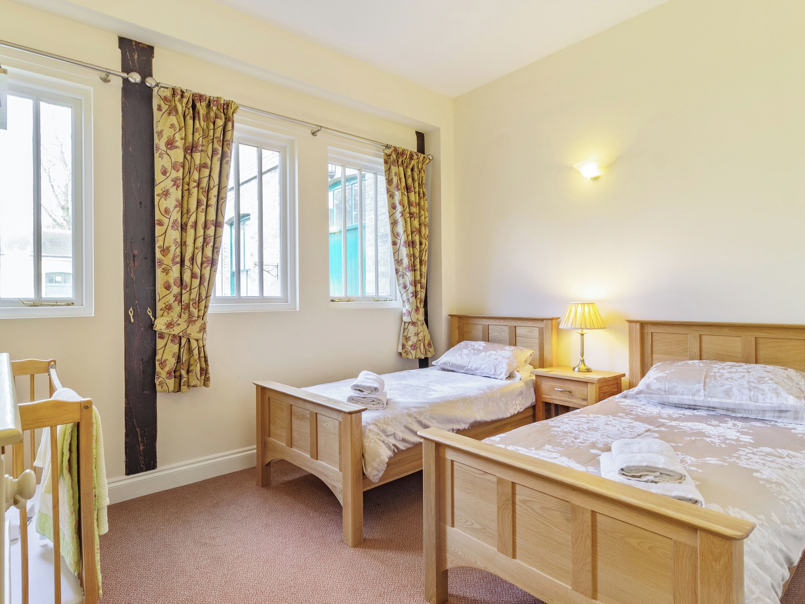 A bedroom with two single beds, a wooden headboard, a bedside table with a lamp, and a window with curtains.