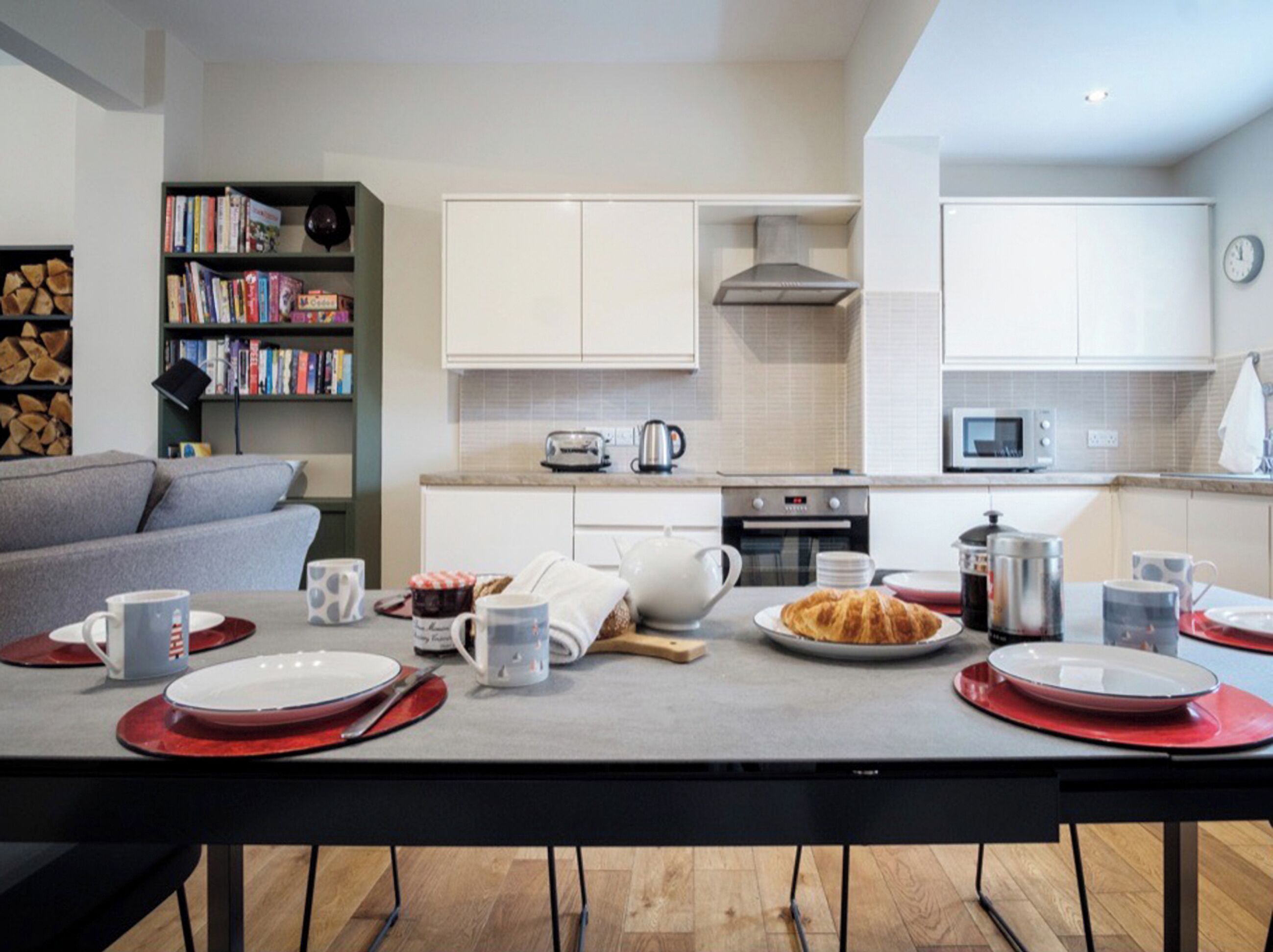 A modern kitchen with white cabinets, a microwave, and a toaster. There is a dining table set with plates, cups, and a croissant.