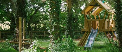 A wooden play structure with a slide in a grassy area surrounded by trees.