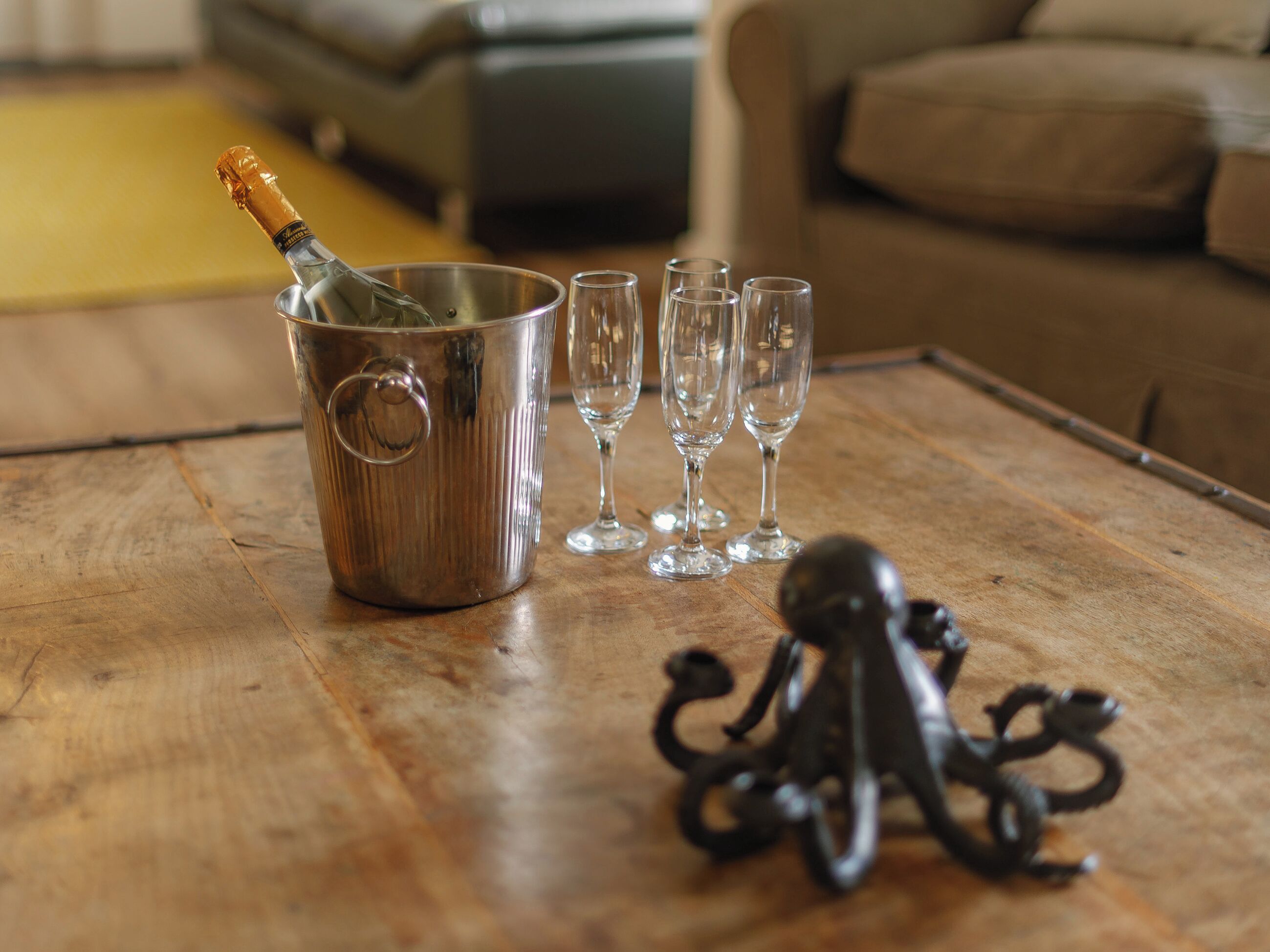 A metal bucket with a bottle of champagne, four empty wine glasses, and a decorative octopus figure on a wooden table.