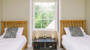Ein Schlafzimmer mit zwei hölzernen Betten, weißer Bettwäsche, einem Fenster mit Blick auf Grünflächen und einem alten Koffer mit Laternen.