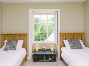 A bedroom with two wooden beds, white bedding, a window with a view of greenery, and a vintage trunk with lanterns. - Corffe House - UKC4970 (Tawstock, near Barnstaple)