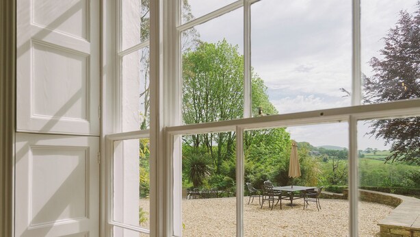 Blick durch ein Fenster auf eine Schotterterrasse mit Gartenmöbeln und Bäumen.