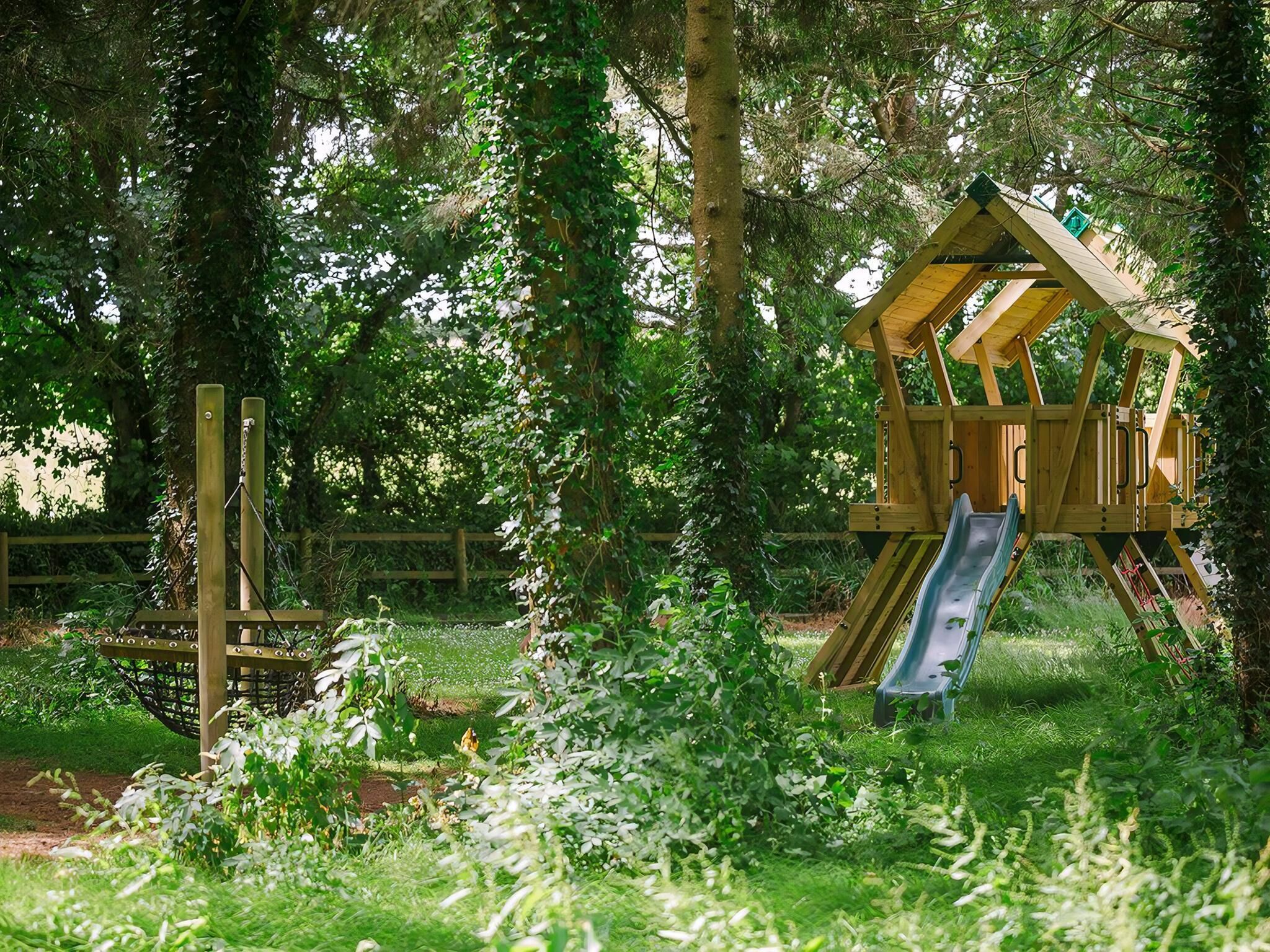 A wooden play structure with a slide in a grassy area surrounded by trees.