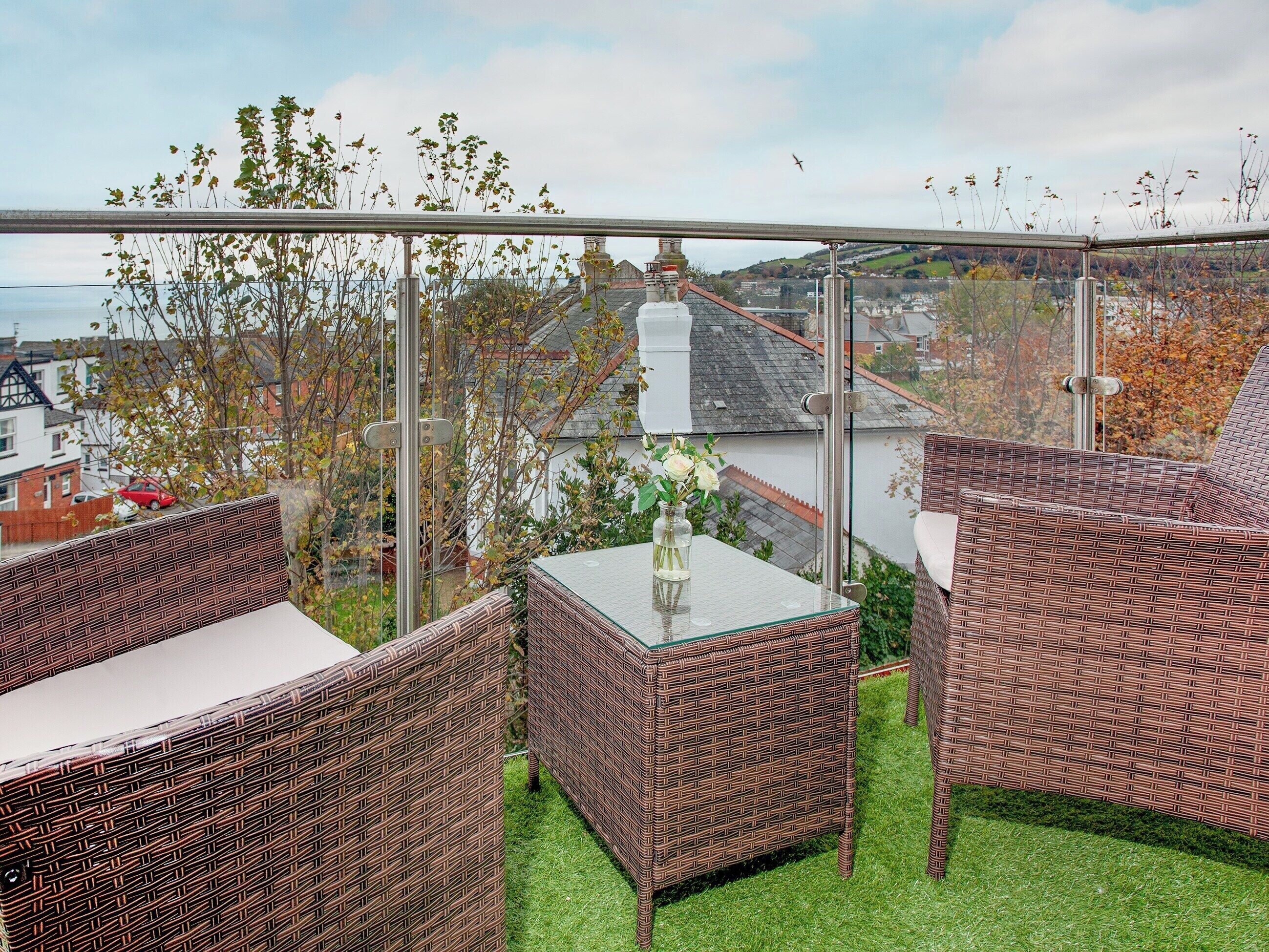 Un balcon agrémenté de meubles en osier et d’une table en verre, donnant sur un quartier résidentiel avec des maisons et des arbres.