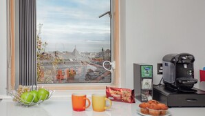 Eine Küche mit Kaffeemaschine, einer Schüssel grüner Äpfel, zwei Tassen, einer Tüte Snacks und Blick auf eine Stadtlandschaft durch ein Fenster.