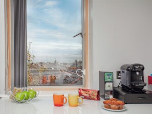 Eine Küche mit Kaffeemaschine, einer Schüssel grüner Äpfel, zwei Tassen, einer Tüte Snacks und Blick auf eine Stadtlandschaft durch ein Fenster.