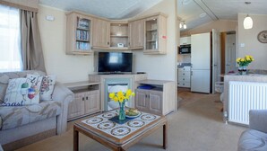 A compact living space with a sofa, a coffee table with a floral vase, a television, and a kitchenette. - 3 bedroom accommodation in Dawlish Warren (Dawlish Warren)