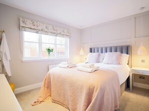 A bedroom with a large bed, a window with a floral valance, a bedside table with a lamp, and a towel rack. - Carn Mor is a beautifully refurbished three bedroom property. (Aviemore)