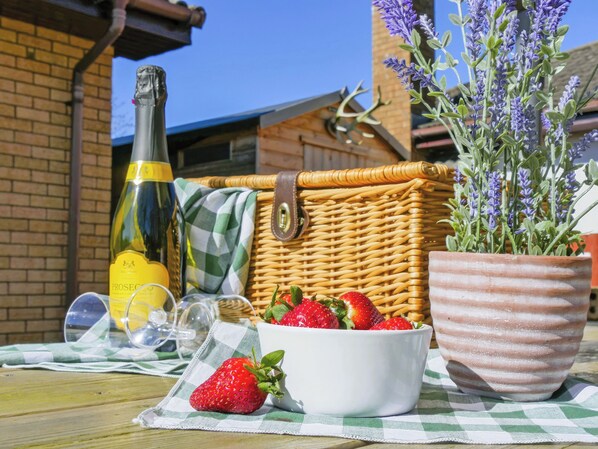 Una cesta de mimbre, una botella de vino, un bol de fresas y una planta de lavanda en maceta sobre un mantel a cuadros.