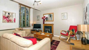 A living room with a beige sofa, a red armchair, a wooden coffee table, a flat-screen TV, a fireplace, and a painting on the wall. - Larrach Beag-UKC5059 (Aviemore)