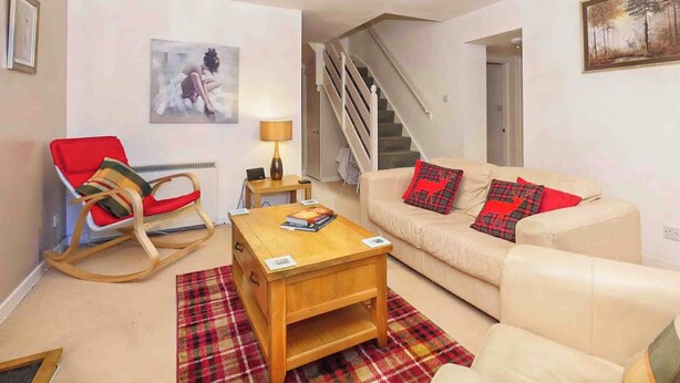 A living room with a beige sofa, red and black plaid rug, wooden coffee table, and a rocking chair. A staircase is visible in the background. - Larrach Beag-UKC5059 (Aviemore)