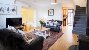 A living room with a black leather sofa, a white coffee table, and a black leather couch. There is a television on a wooden stand, a framed picture on the wall, and a staircase leading to the upper floor. - Ruby Cottage-UKC5064 (Aviemore)