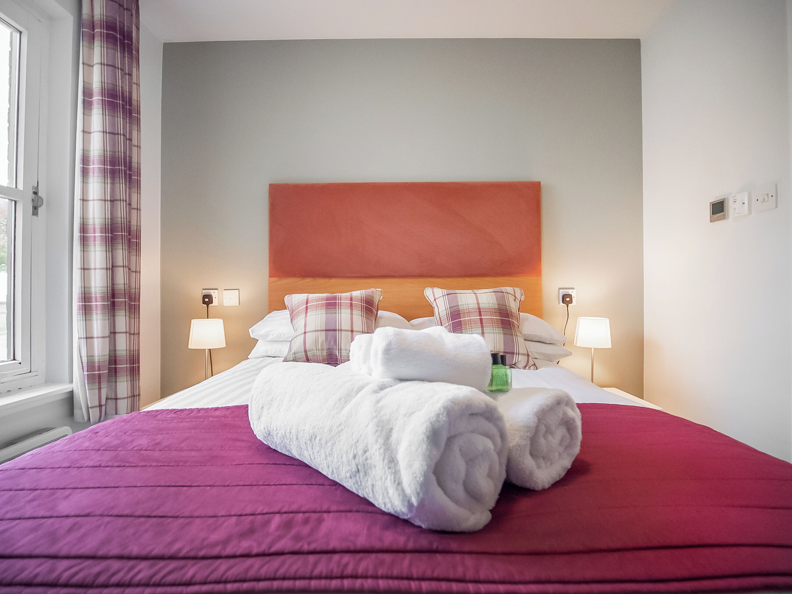Una cama bien hecha con una colcha roja, dos almohadas y una toalla blanca enrollada.